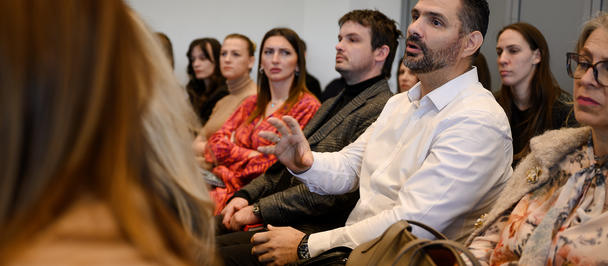 Back of a person with red hair facing a conference-room audience; a man in white raises his hand.