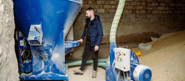 Photograph of a man inspecting a large blue industrial hopper beside a beige material pile.