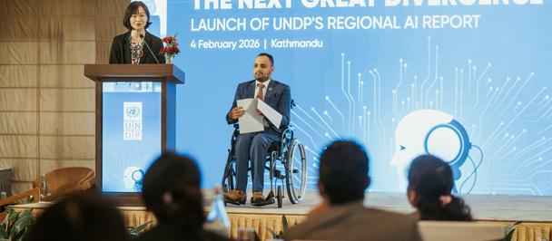 Speaker at podium and a wheelchair user on stage at a conference with a blue backdrop.