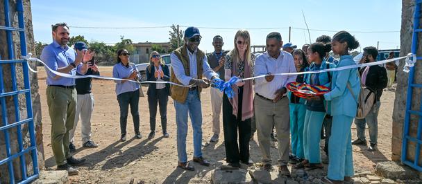 Group of people at a groundbreaking ceremony, standing behind a blue ribbon across a dirt site.