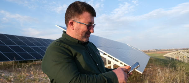 Person in a green jacket outdoors beside solar panels, holding a rectangular panel.