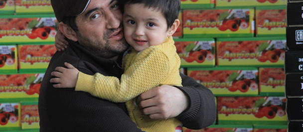 Two people hug, one wearing black and the other yellow, in front of a wall of colorful boxes.