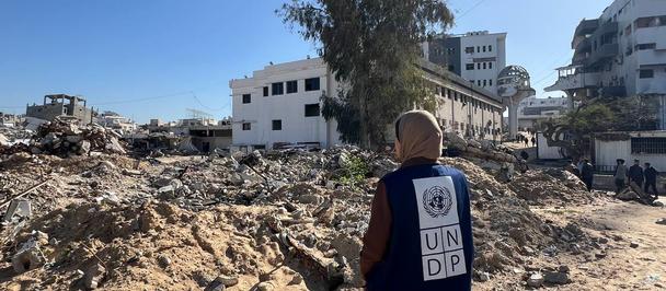 A person in a red UN vest stands amid rubble and damaged buildings under a clear blue sky.