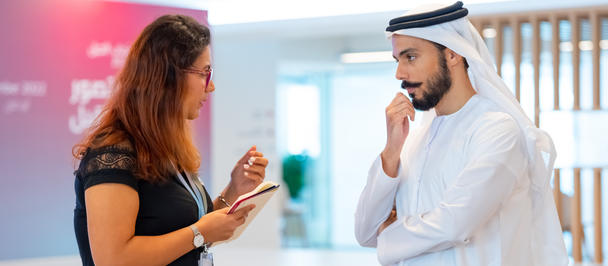 Woman in black top shows her phone to a man in a white kandura in a bright, modern lobby.