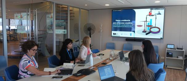 Masked team sits around a conference table in a modern office, slide on screen at front.