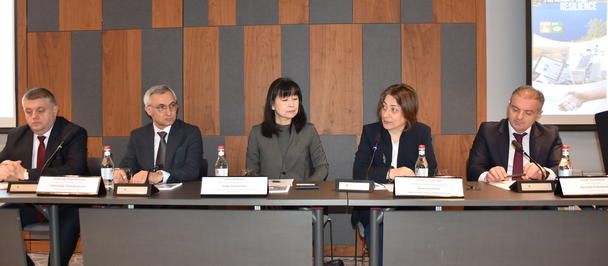 Five panelists sit at a conference table with nameplates and microphones.