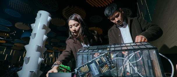 Two people examine a transparent control panel on a lab rig with a green turf surface.