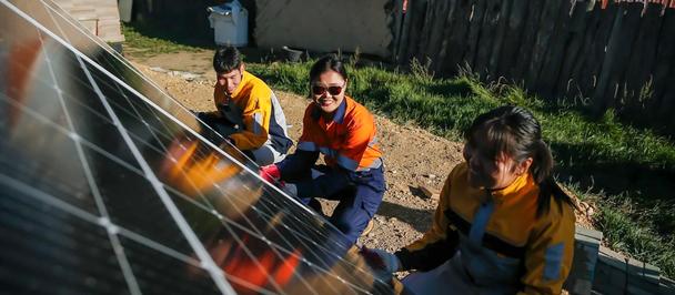 Three workers in orange safety vests kneel beside solar panels in a yard.