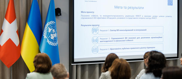 Audience in a conference room watches a slide with Swiss, Ukrainian, and UN flags beside screen.