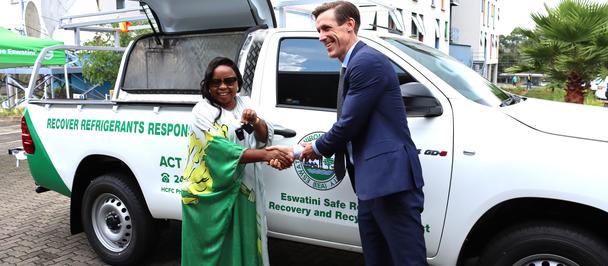 A man in a dark suit hands a key to a woman in a green sari beside a white branded van.