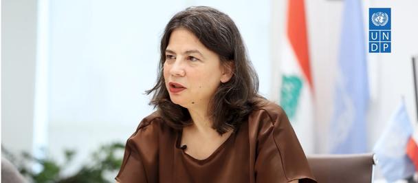 Woman with blurred face in a brown blouse seated indoors, flags in the background.