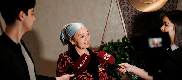 Three people in a room; right interviewer, left man, center red-black outfit with blue headscarf.