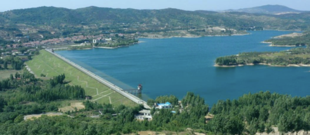 Photograph of a long bridge crossing a blue lake, with green hills and distant mountains.
