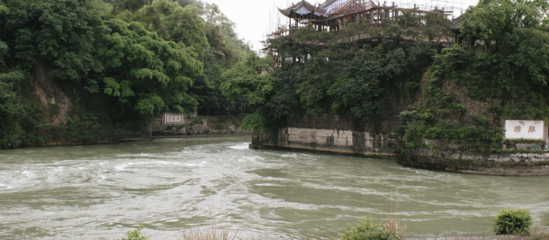 River flows between green banks, with a traditional building atop a rocky hill.