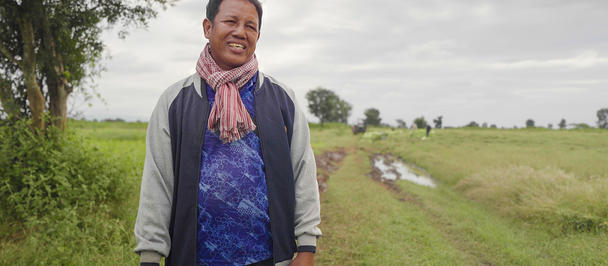 Photograph of a farmer standing on a dirt path in a grassy rural field.