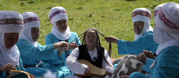 Turquoise-clad women with headscarves sit in grass circle around a girl in a black-and-white dress.