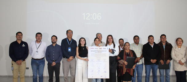 Group of people on stage posing with a large white placard, 12:06 projection behind them.