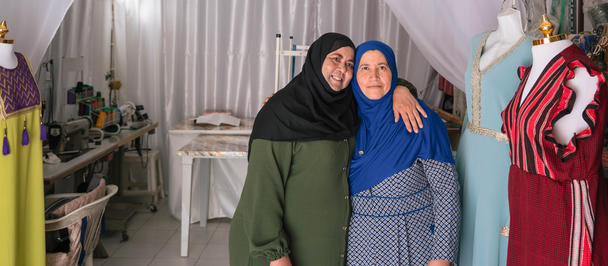 Two women in hijabs pose in a sewing workshop surrounded by mannequins in colorful gowns.