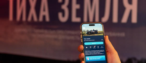 Hand holding a smartphone in front of a large sign with Cyrillic text ТИХА ЗЕМЛЯ in the background.