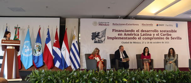 Panel discussion on stage with five speakers, podium, and flags beside a Spanish banner.