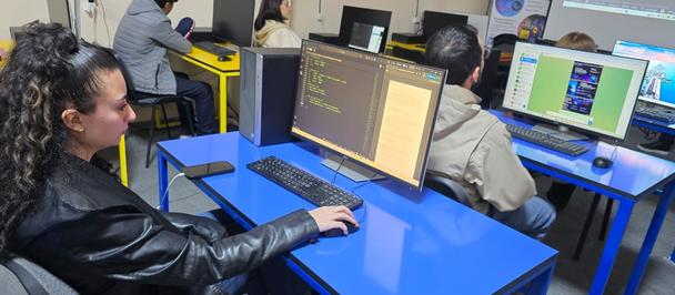 Photograph of a computer lab with blue desks; a woman sits at the left using a keyboard.