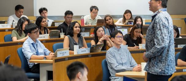 Photograph: lecturer in a patterned shirt standing at the front of a classroom full of students.