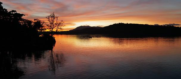 Sunset over calm lake with silhouettes of trees and distant hills, reflecting orange sky.