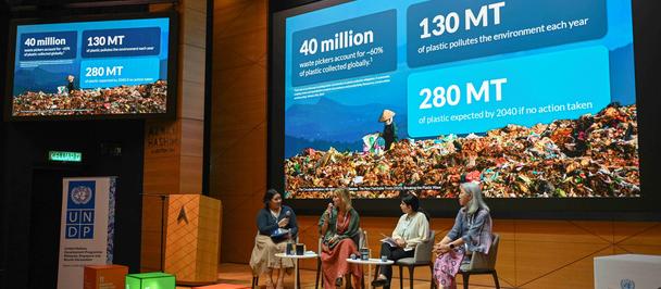 Photograph of a panel on a lit stage with speakers and large blue presentation screens.
