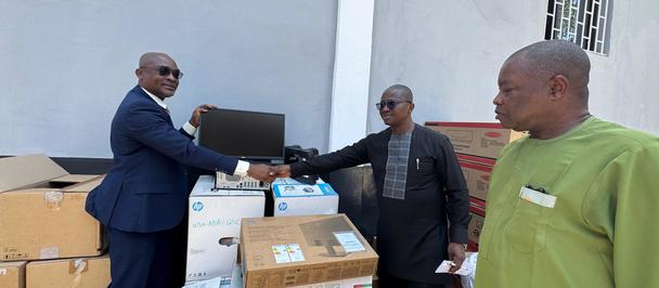 Three men in business attire outside a white building, handing a black folder near donated boxes.