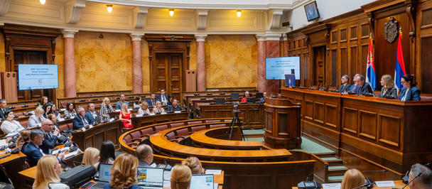 Photo of a large wood-paneled parliamentary chamber with a speaker at a podium and an audience.