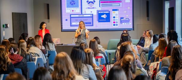 Photograph of a conference room with a presenter onstage, seated audience, and slides projected.