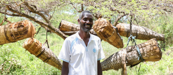 Smiling man in white shirt carries a large bundle of logs on his shoulders outdoors.