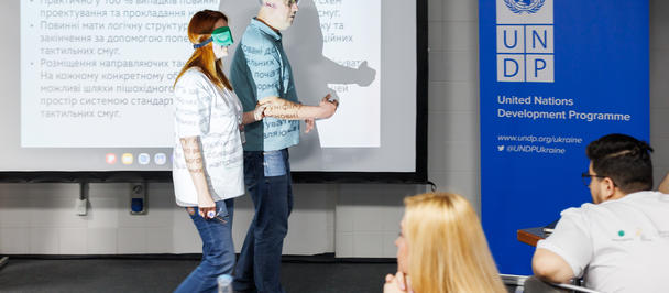 People in masks walk at the front of a conference room with a UNDP banner and projector screen.