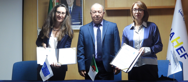 Three people stand behind a table; two hold documents at a formal ceremony, flags in background.