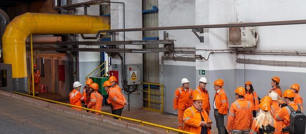 Group of workers in orange safety outfits with hard hats near yellow pipes at an industrial plant.