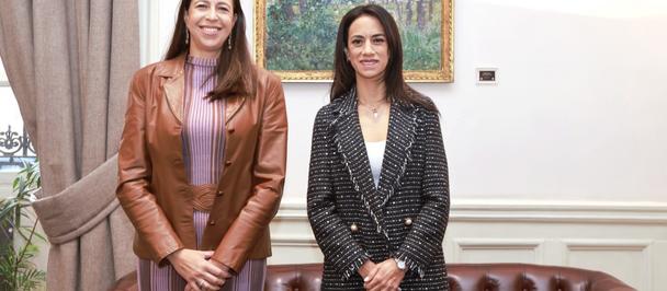 Photograph: two women in business attire stand in a living room with a brown leather sofa and framed painting.