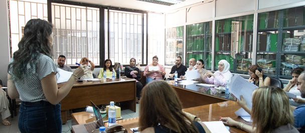 Photograph of a woman presenting to a long table full of attendees in a bright meeting room.