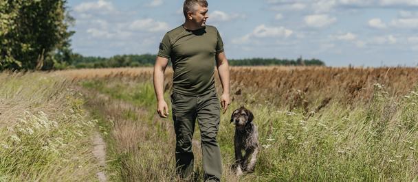 Photo: Man in olive shirt walks along a grassy path with a dog in a field, blue sky.