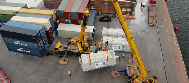Aerial view of a port with stacked shipping containers and a yellow crane lifting a white object.