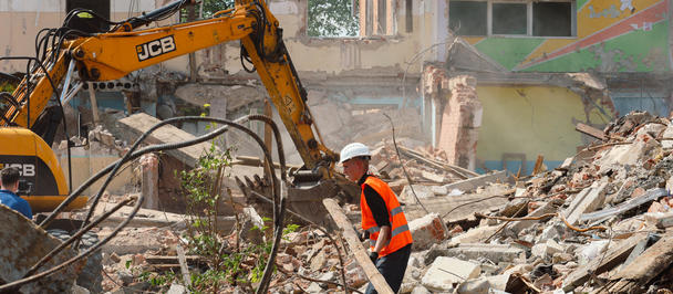 Yellow excavator and a worker in an orange safety vest amid rubble and damaged buildings.
