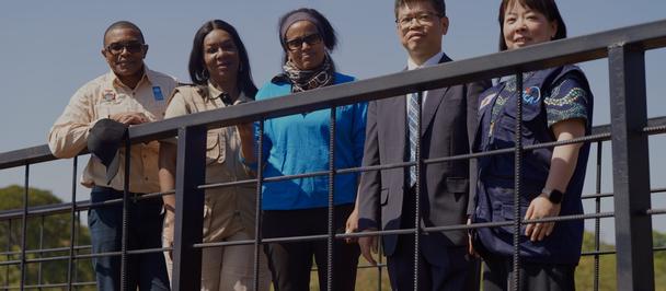Five adults posing behind a railing outdoors, smiling, against a blue sky.