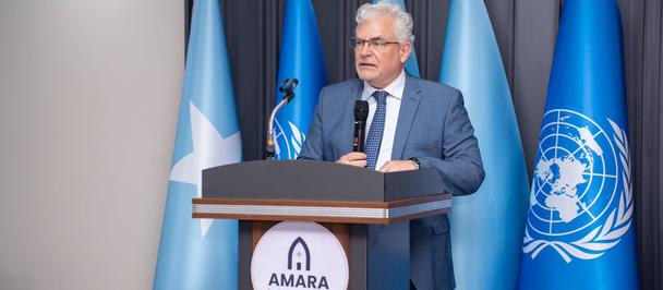 Man in a blue suit with gray hair delivers a speech at a podium, flags behind him.