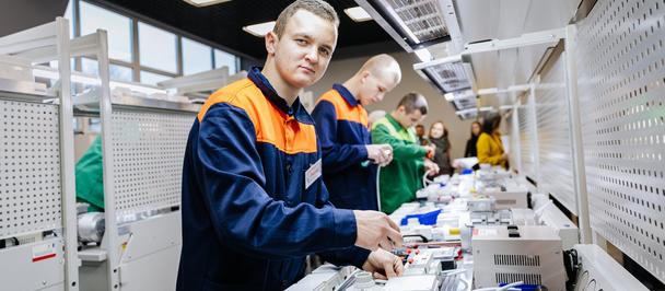 Factory workers in blue and orange uniforms assemble parts at a long production line.