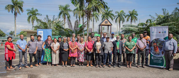 Group photo of people posing outdoors with palm trees and banners in a sunny coastal setting.