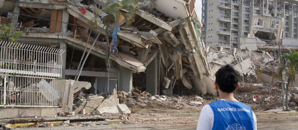 Person in a blue safety vest surveys a collapsed multi-story building and rubble.