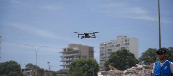 Drone over a busy street with a man in a blue vest; urban buildings and rubble in the background.