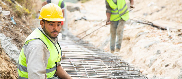 Construction worker in a yellow hard hat and safety vest sits beside a rebar trench.