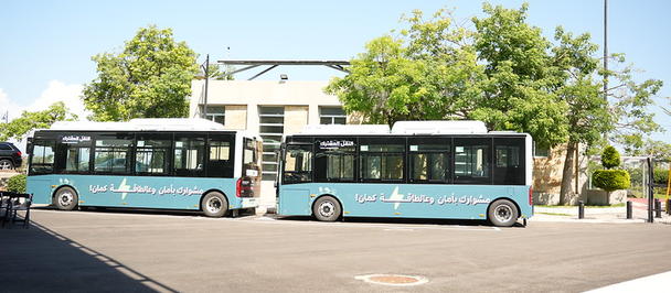 Two blue transit buses parked in a sunny lot beside a building with trees.