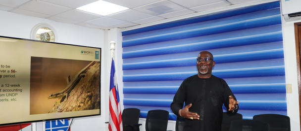 Photo: man in dark clothing stands behind a conference desk; blue striped wall, flags, plaque.