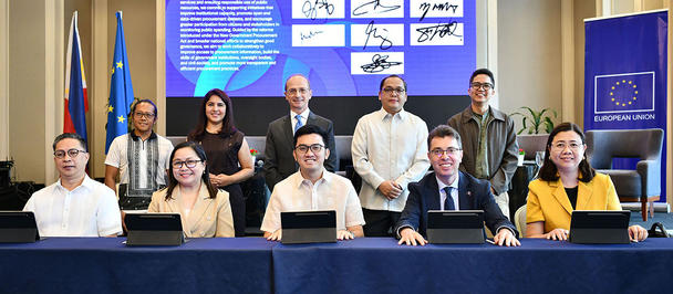 Group photo of professionals at a conference panel, seated behind a blue table with a large screen.